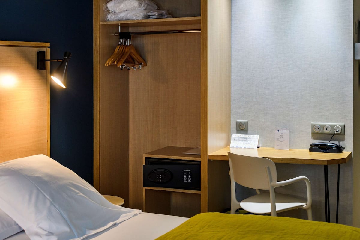 A hotel room with a bed, a desk, a chair, a wardrobe, and a desk lamp.