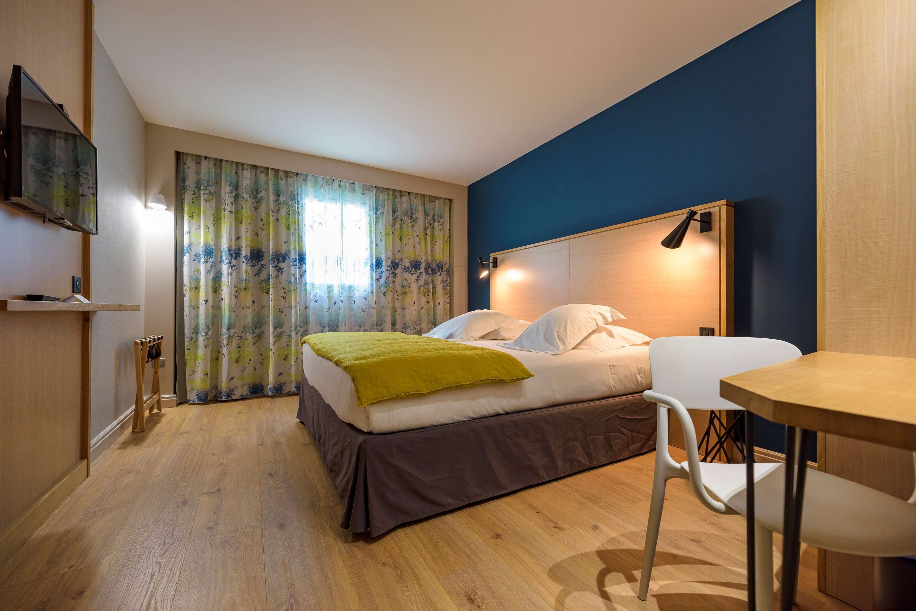Een slaapkamer met een bed, een bureau, een stoel, een televisie en een raam met gordijnen.