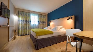 Un dormitorio con cama, escritorio, silla, televisor y ventana con cortinas.
