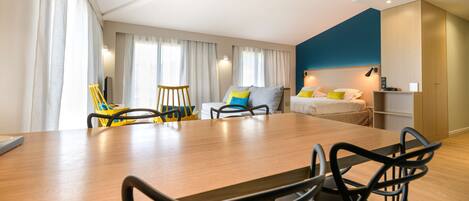 Une chambre d’hôtel moderne avec une table à manger en bois, des chaises noires, un lit avec une tête de lit bleue et une petite kitchenette.
