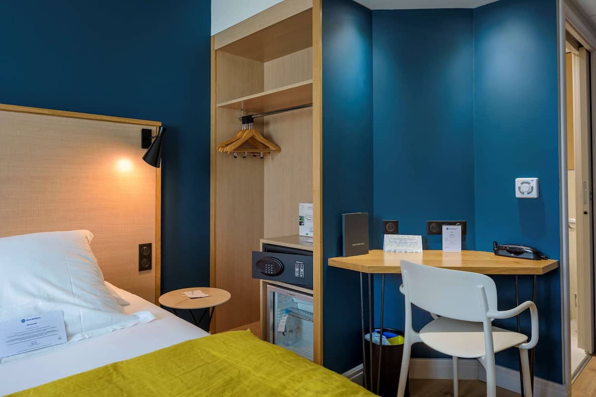 A hotel room with a bed, a desk, a chair, a mini-fridge, a coat rack, and a small table with a lamp.