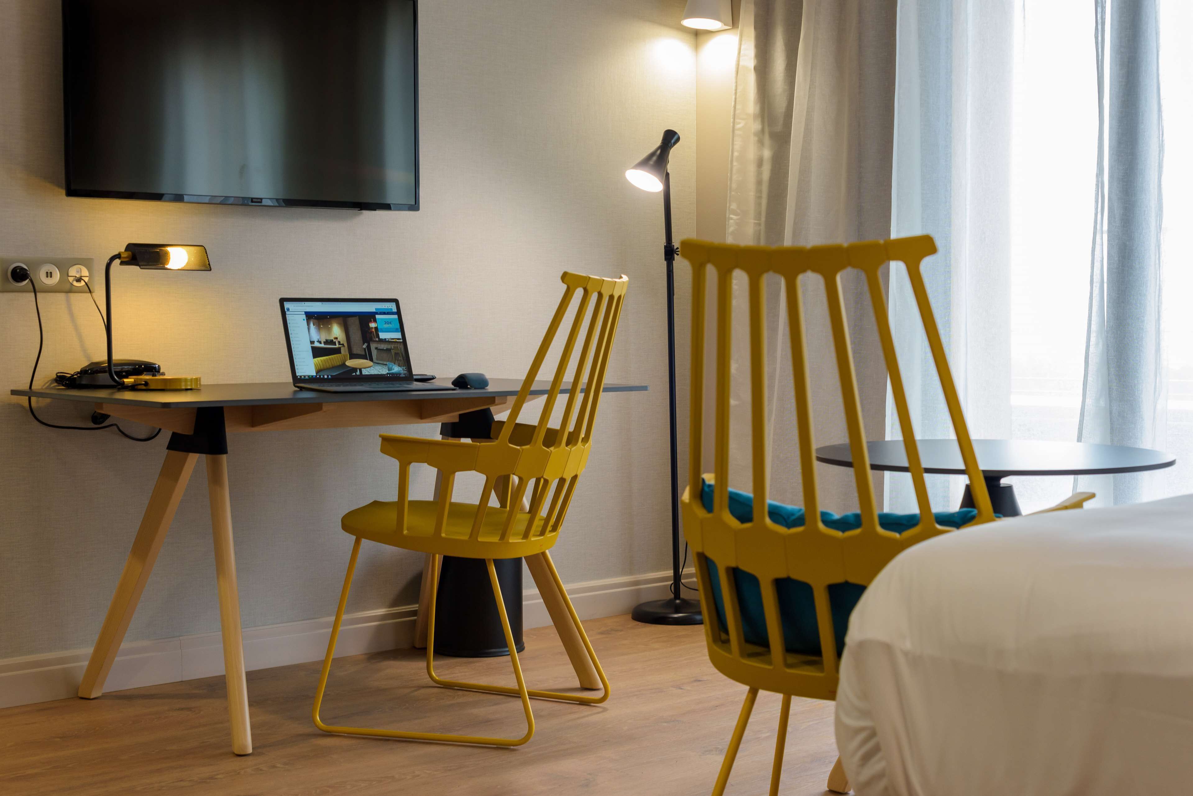 Una camera d'albergo con una scrivania, un portatile, una sedia gialla, un letto e una TV a muro.