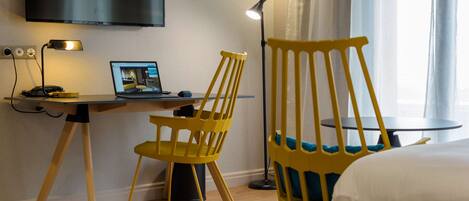 A hotel room with a desk, a laptop, a yellow chair, a bed, and a TV mounted on the wall.