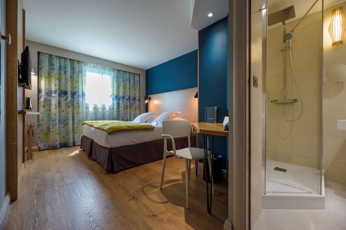 A hotel room with a bed, a desk, a chair, a shower, and a television.