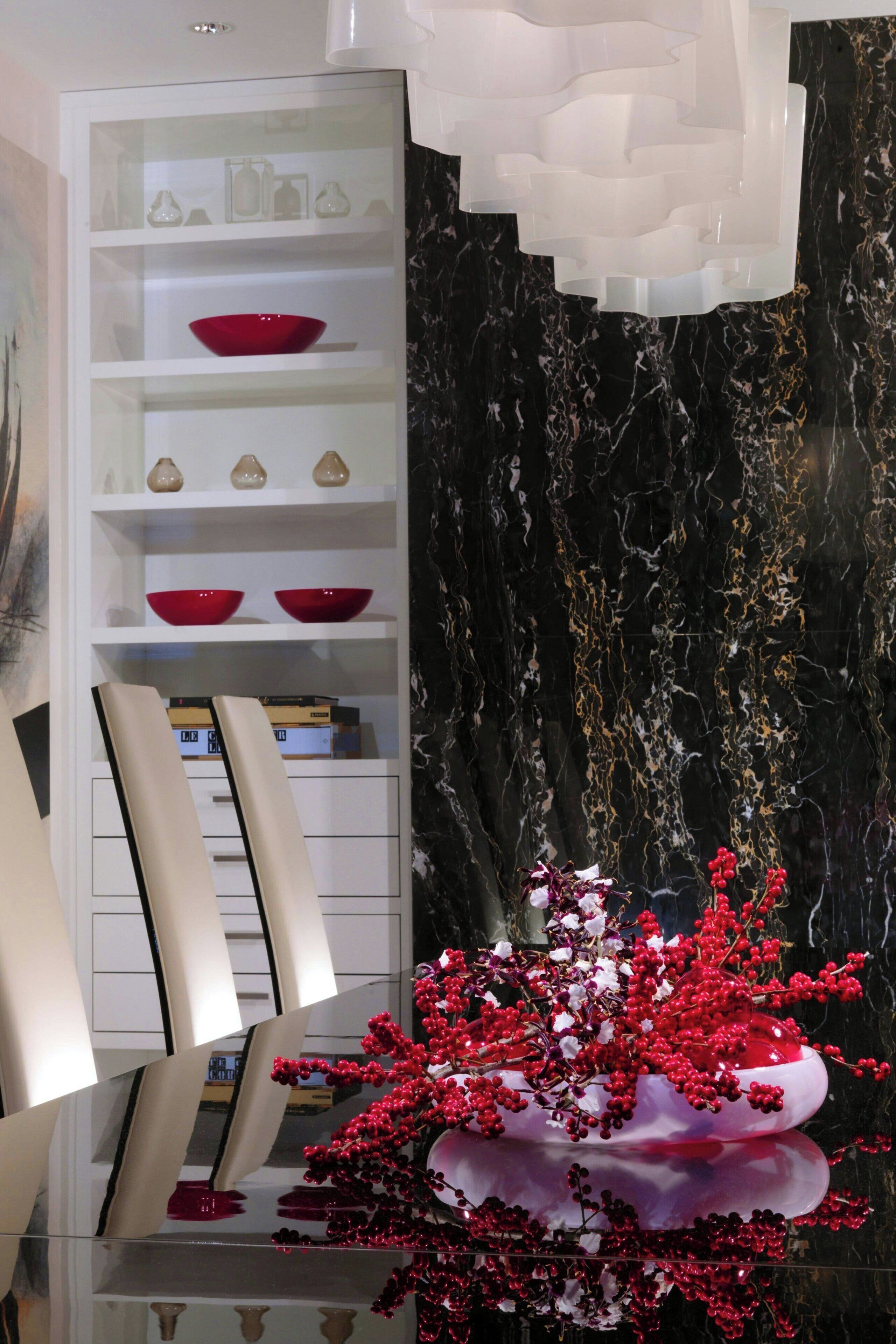A modern interior with a white shelving unit displaying red bowls and decorative items, a reflective glass table, and a black marble wall with gold veins.