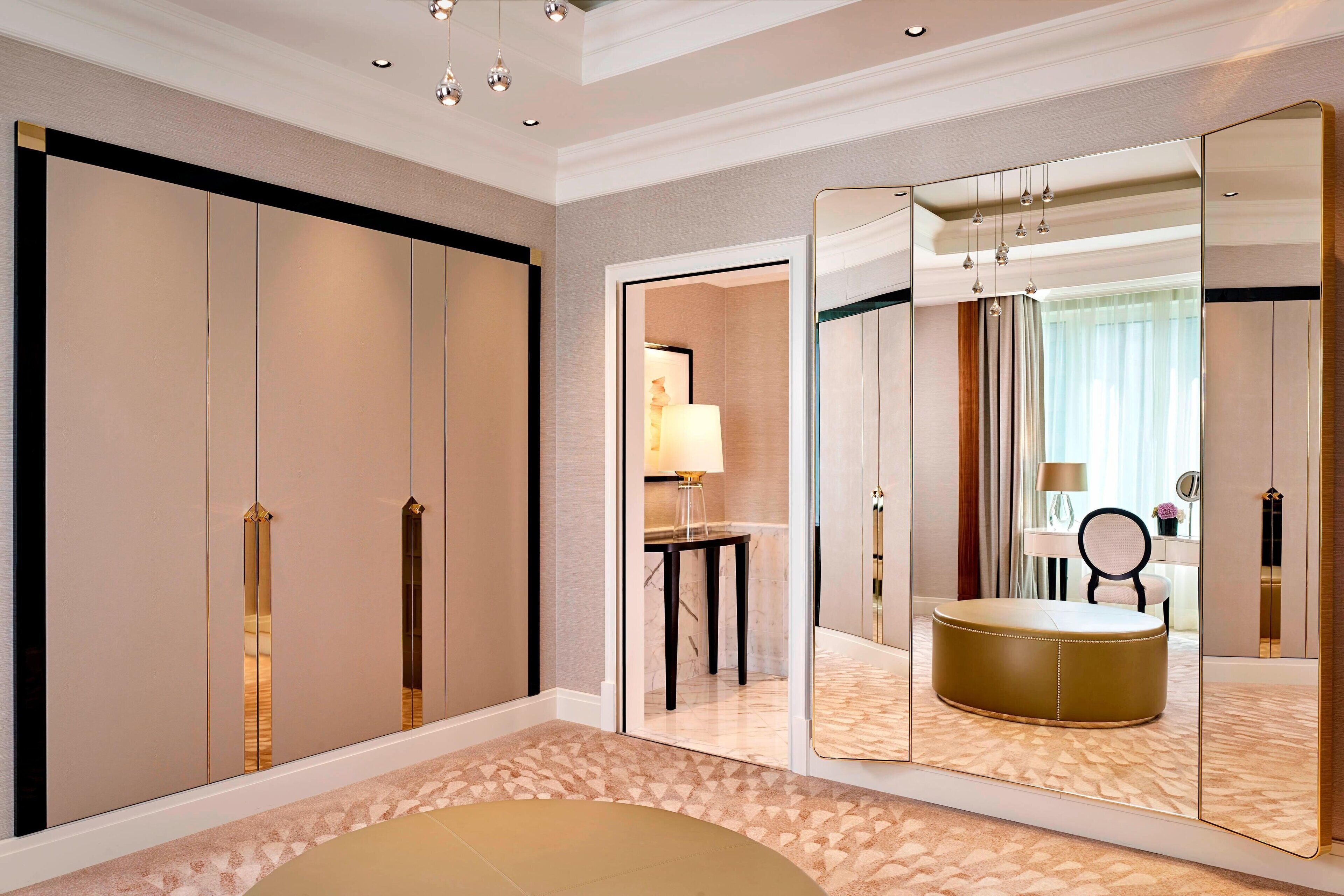 A hotel room with a large mirror, a round ottoman, a small table with a lamp, and a view of a bathroom through an open door.