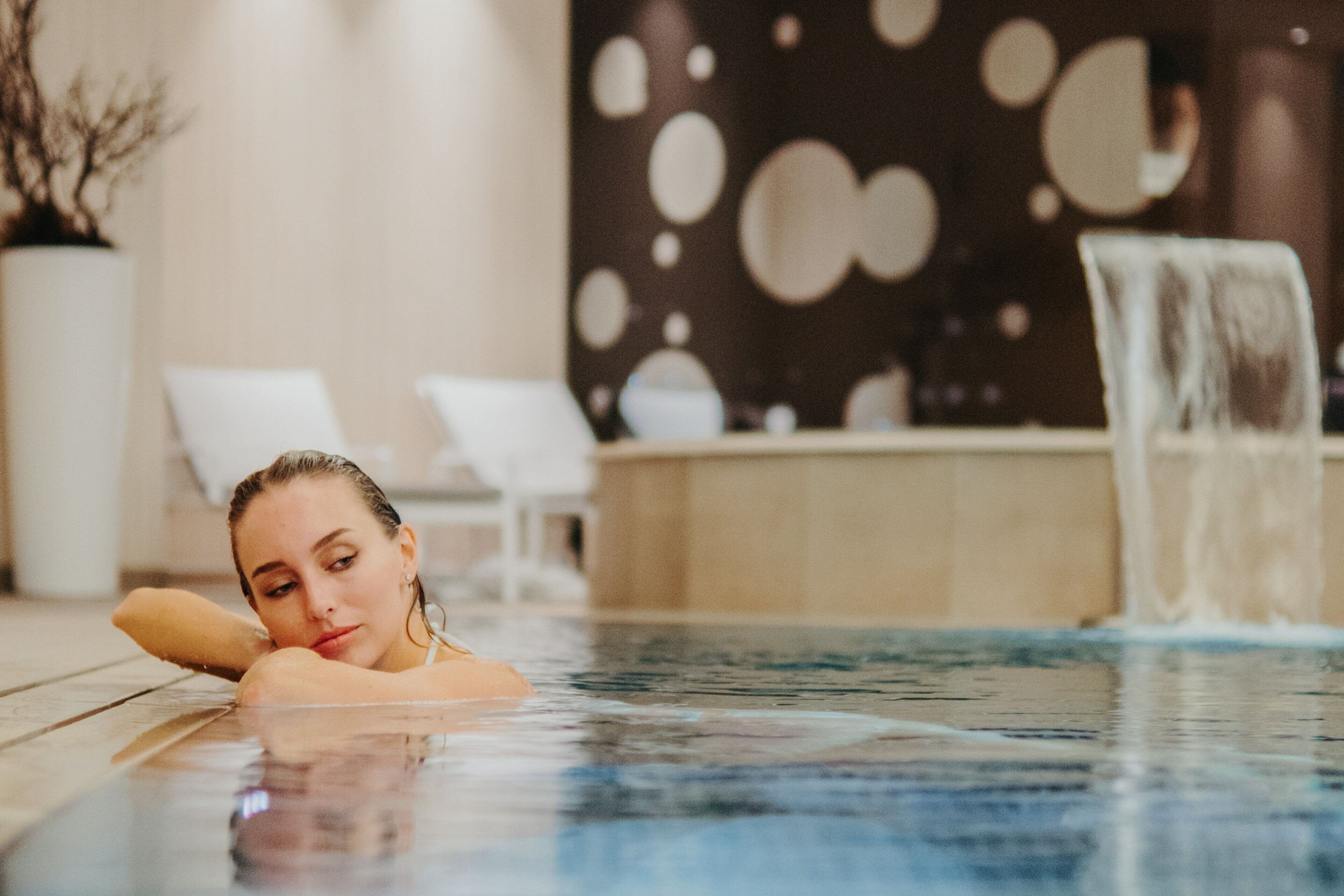 A person in a pool with a blurred background featuring a wall with circular patterns and a potted plant.