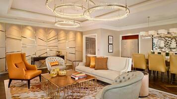 A spacious living room with a white sofa, a tufted armchair, and a wooden coffee table. There are two large chandeliers and a dining area with yellow chairs.