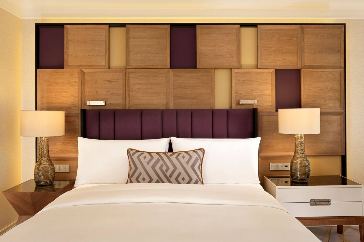 Habitación de hotel con cabecera de madera, dos mesitas de noche con lámparas, cama con ropa blanca y un cojín decorativo estampado.