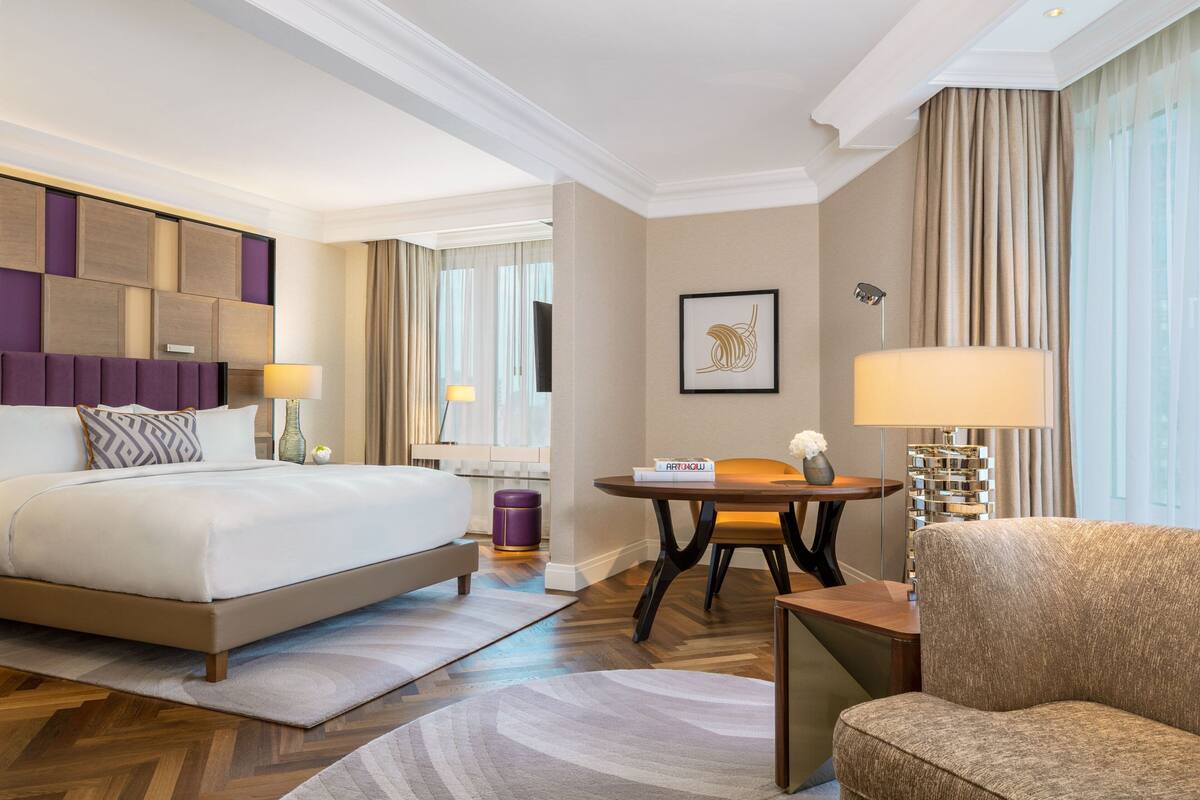 Habitación de hotel con una cama grande, un espacio para sentarse con una mesa y una silla, y vista a la ciudad por la ventana.