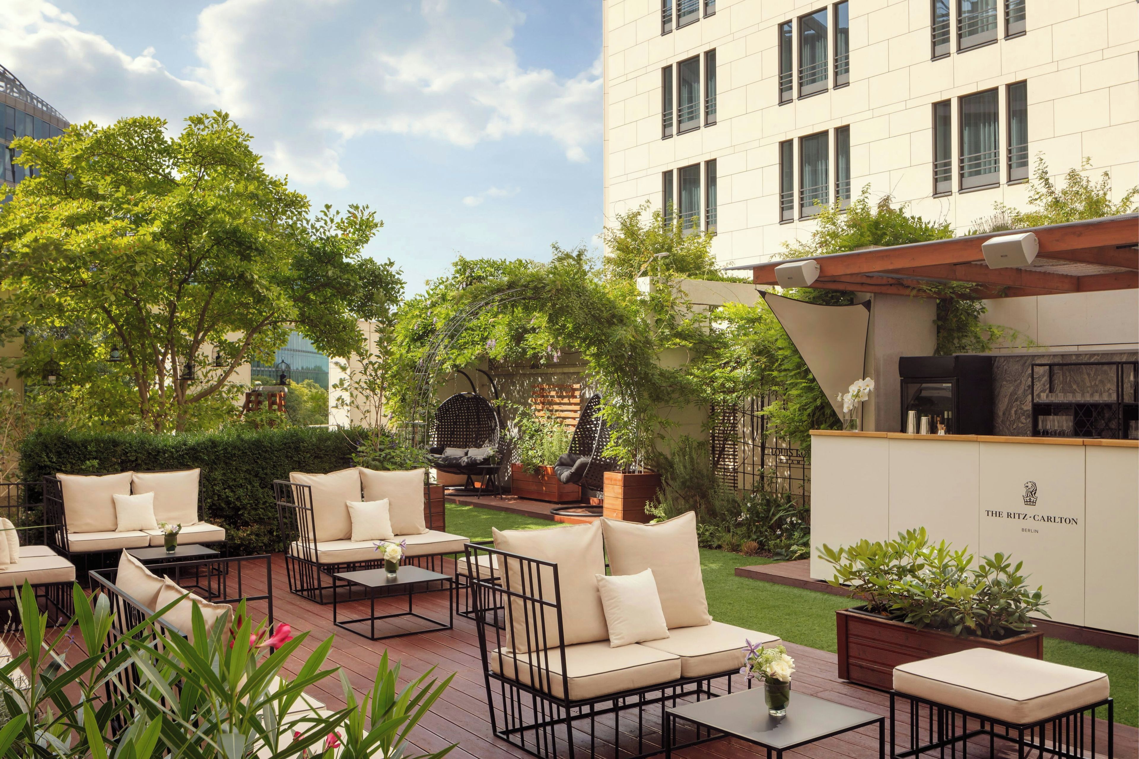 Un área de estar al aire libre con varios sofás y mesas, rodeada de follaje y un edificio con un letrero que dice 'The Ritz-Carlton Berlin'.