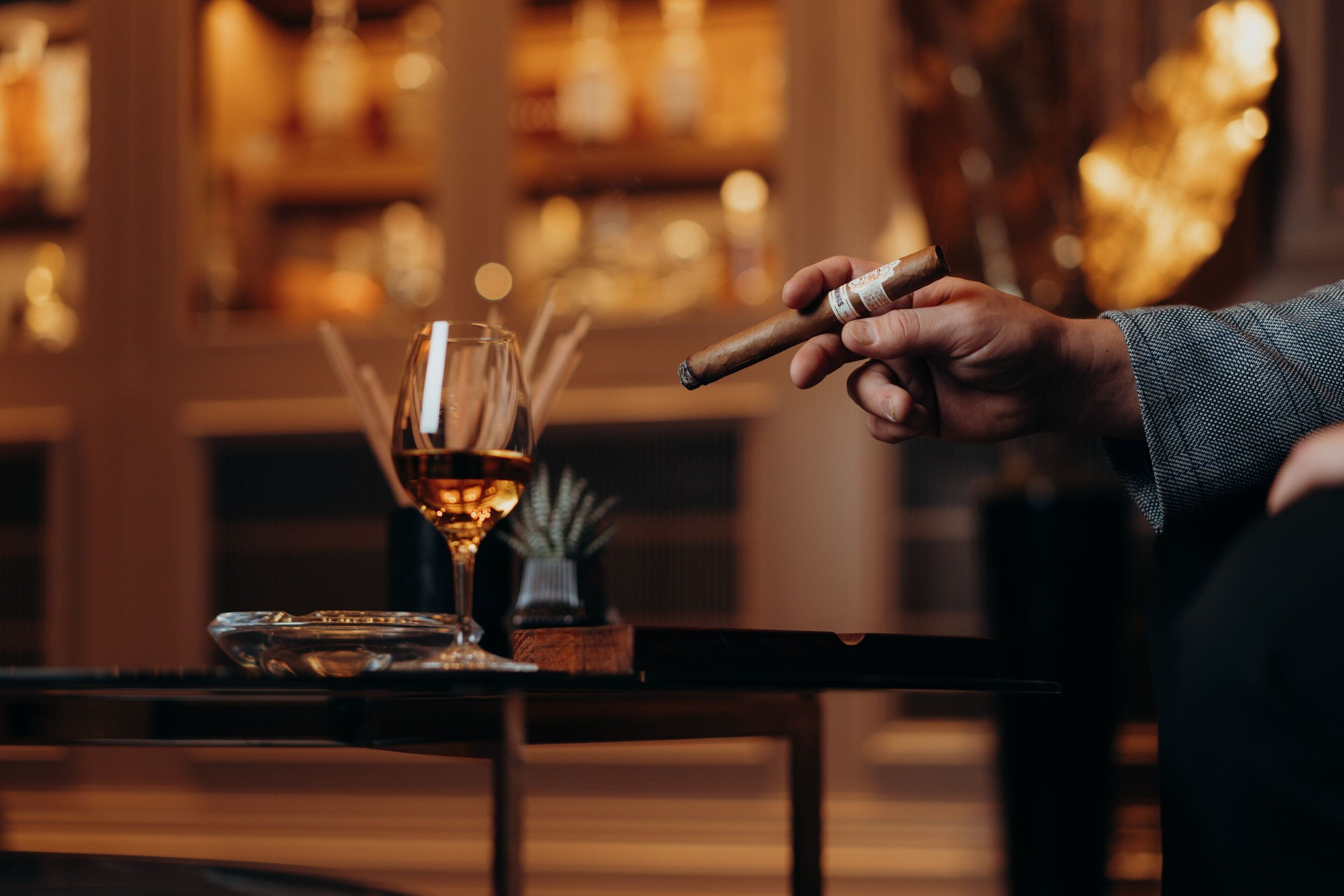 A person holding a cigar, a glass of whiskey, and a cigar box on a table.