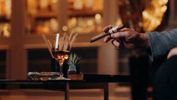 A person holding a cigar, a glass of whiskey, and a cigar box on a table.