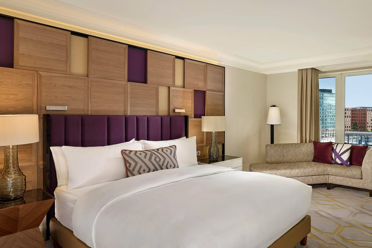 Habitación de hotel con una cama grande, un sofá, una mesita de noche con lámpara y vistas a la ciudad por la ventana.