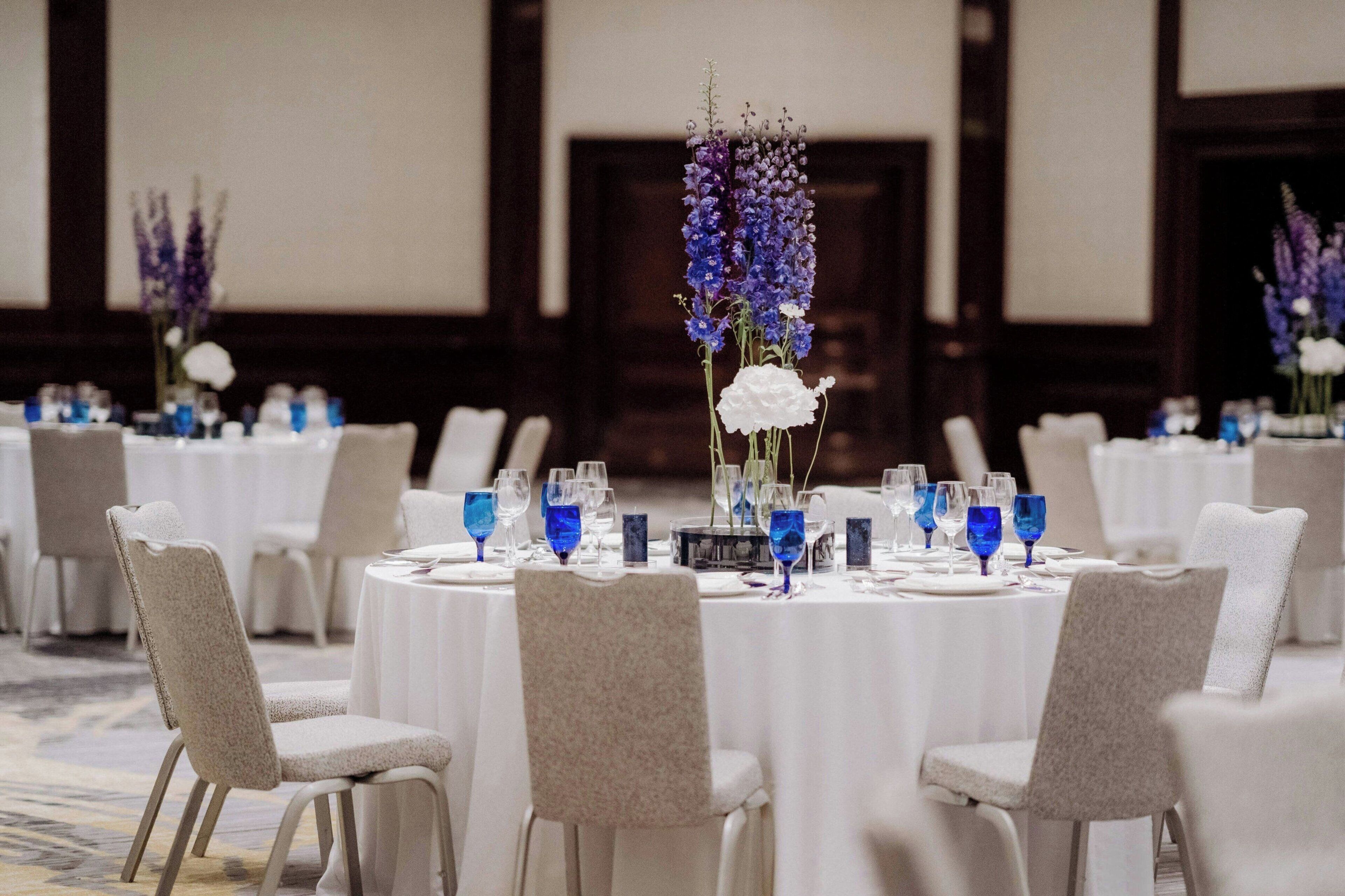A banquet hall with round tables set for dining, featuring white tablecloths and blue glassware.