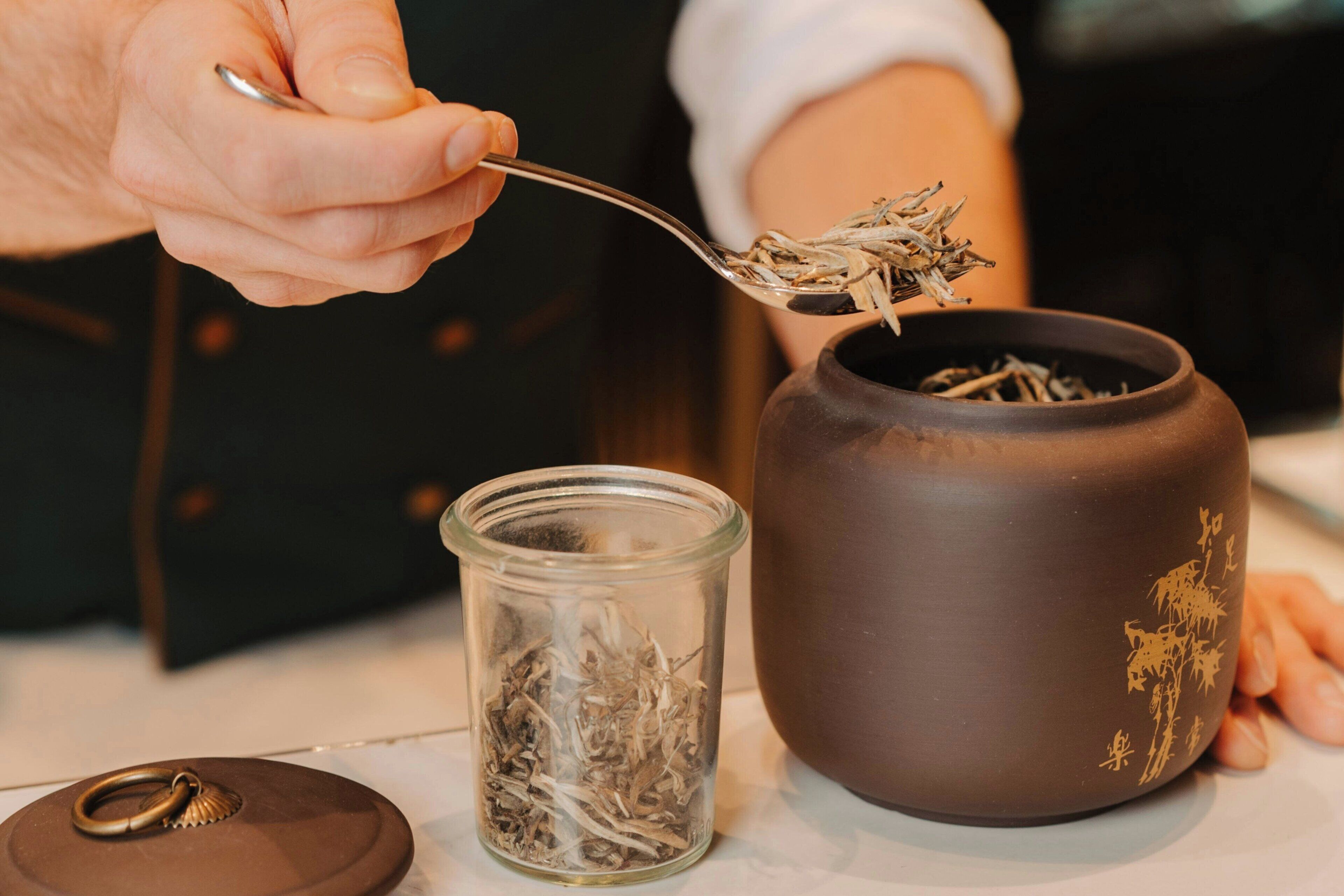 A person holding a spoon with tea leaves above a ceramic pot.