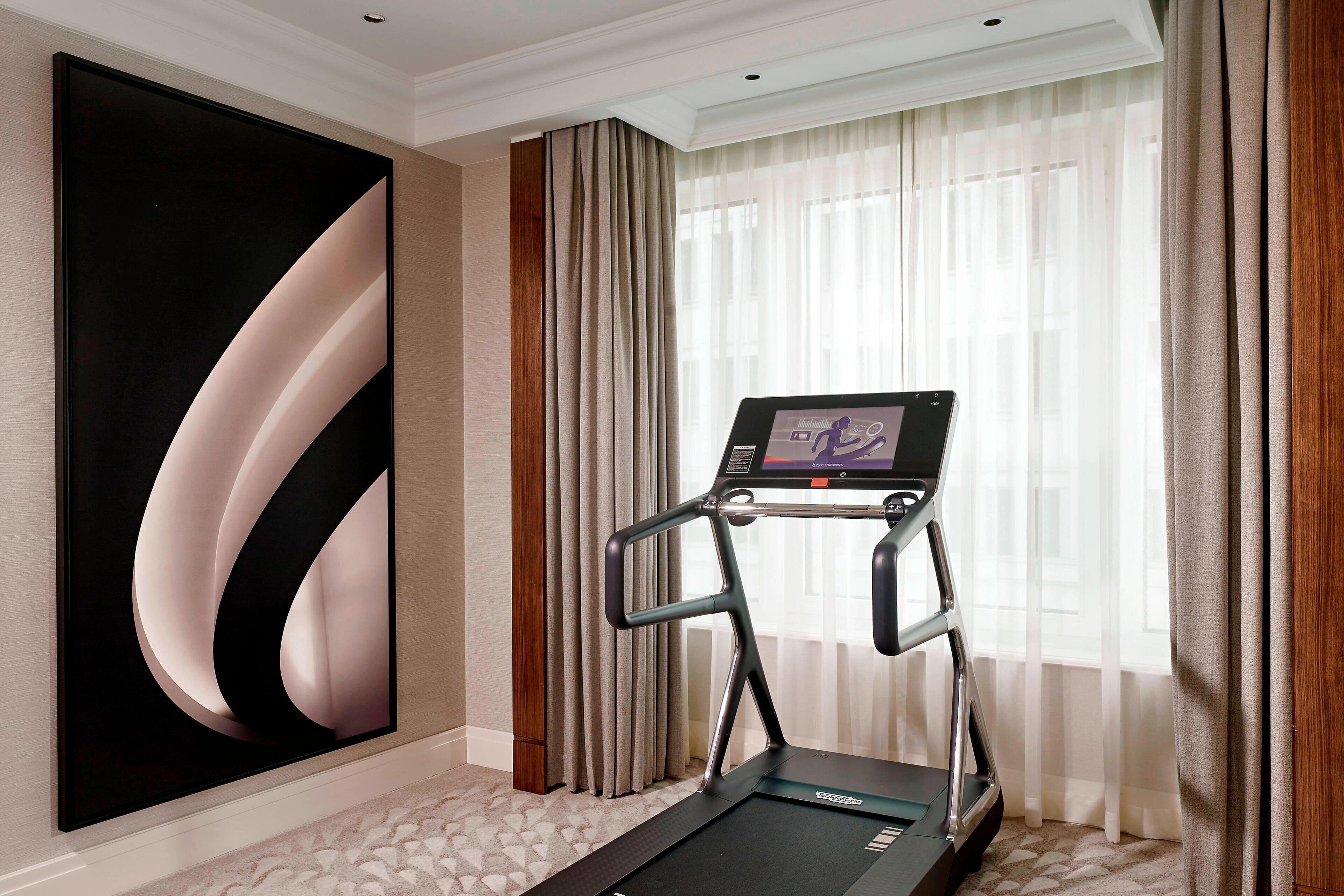 A treadmill in a hotel room with a large abstract painting on the wall, a window with sheer curtains, and a carpeted floor.