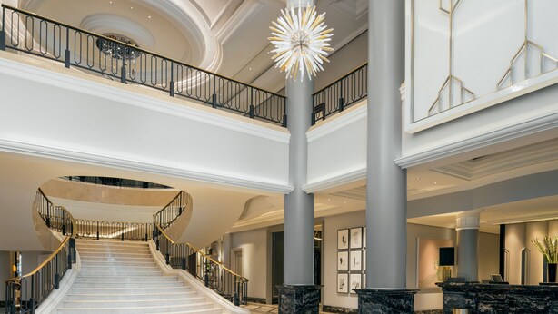 A grand hotel lobby with a central staircase, marble flooring, and a modern chandelier.