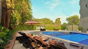 A poolside area with lounge chairs and umbrellas, surrounded by greenery and a clear sky.