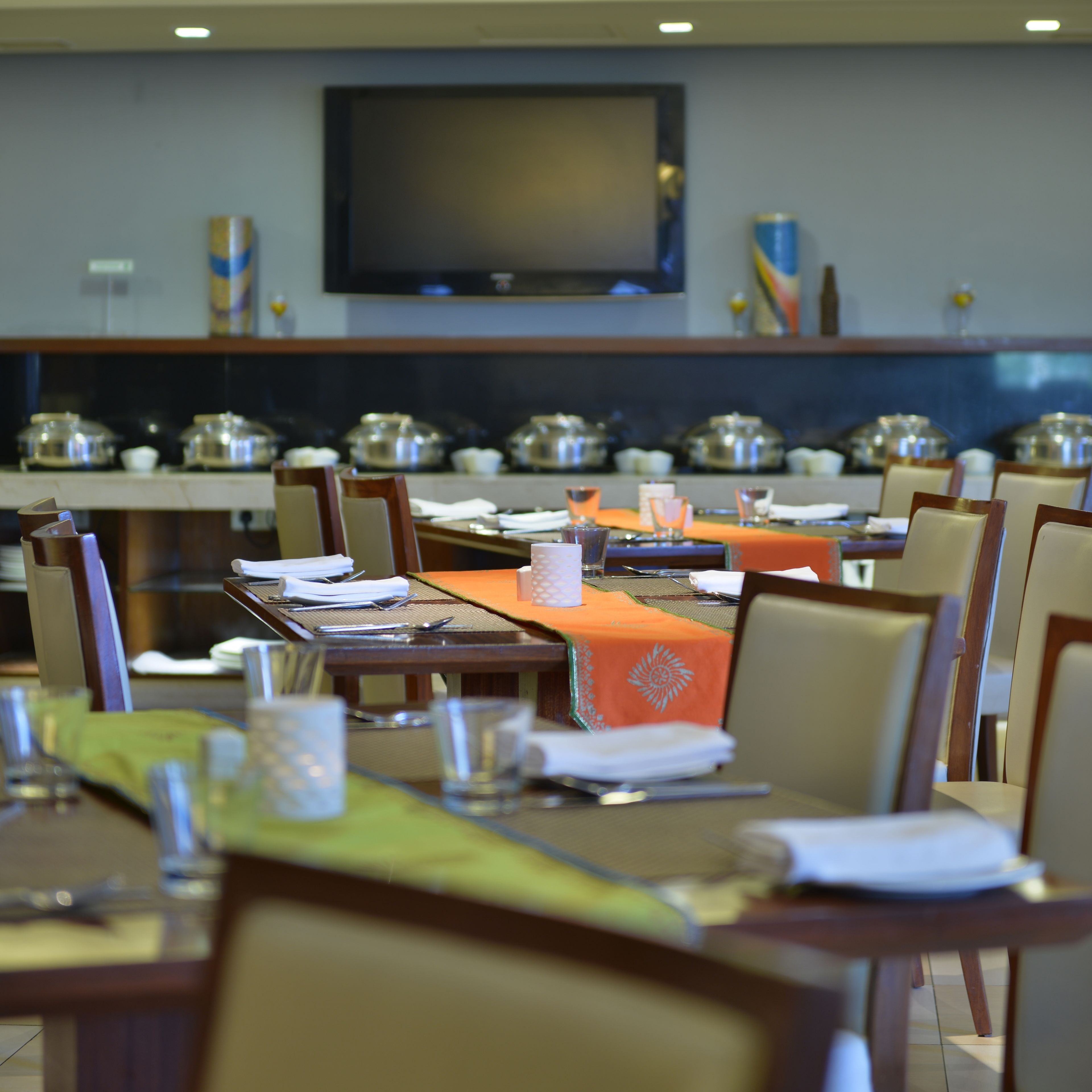 Un comedor con mesas dispuestas para comer, un televisor montado en la pared y servicio de buffet al fondo.