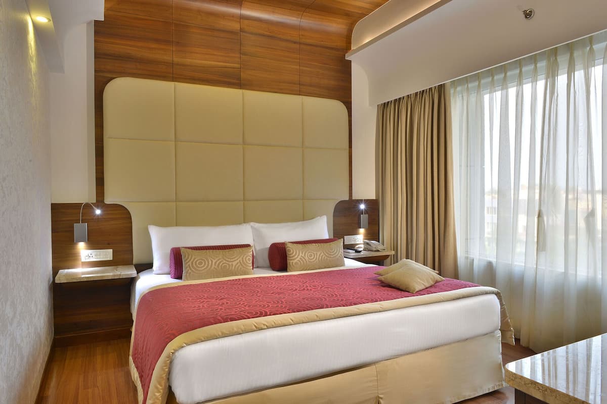 Une chambre d’hôtel avec un grand lit, une tête de lit en bois, des tables de chevet avec des lampes et une vue sur la ville à travers de fines rideaux.