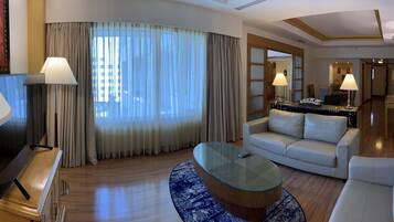 A hotel room with a curved glass coffee table, a sofa, a dining area with a table and chairs, and a large window with curtains.