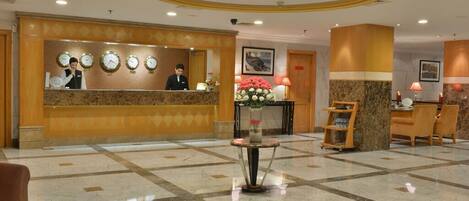 A hotel lobby with a reception desk, clocks, a vase with flowers, and a seating area.