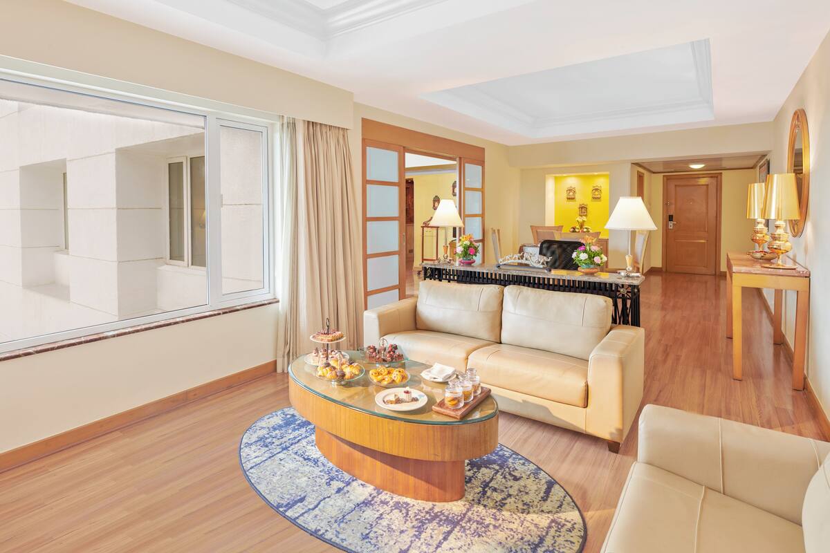 Une chambre d’hôtel spacieuse dotée d’une grande fenêtre, d’une table basse ronde avec des collations, d’un canapé et d’un coin repas avec une table et des chaises.