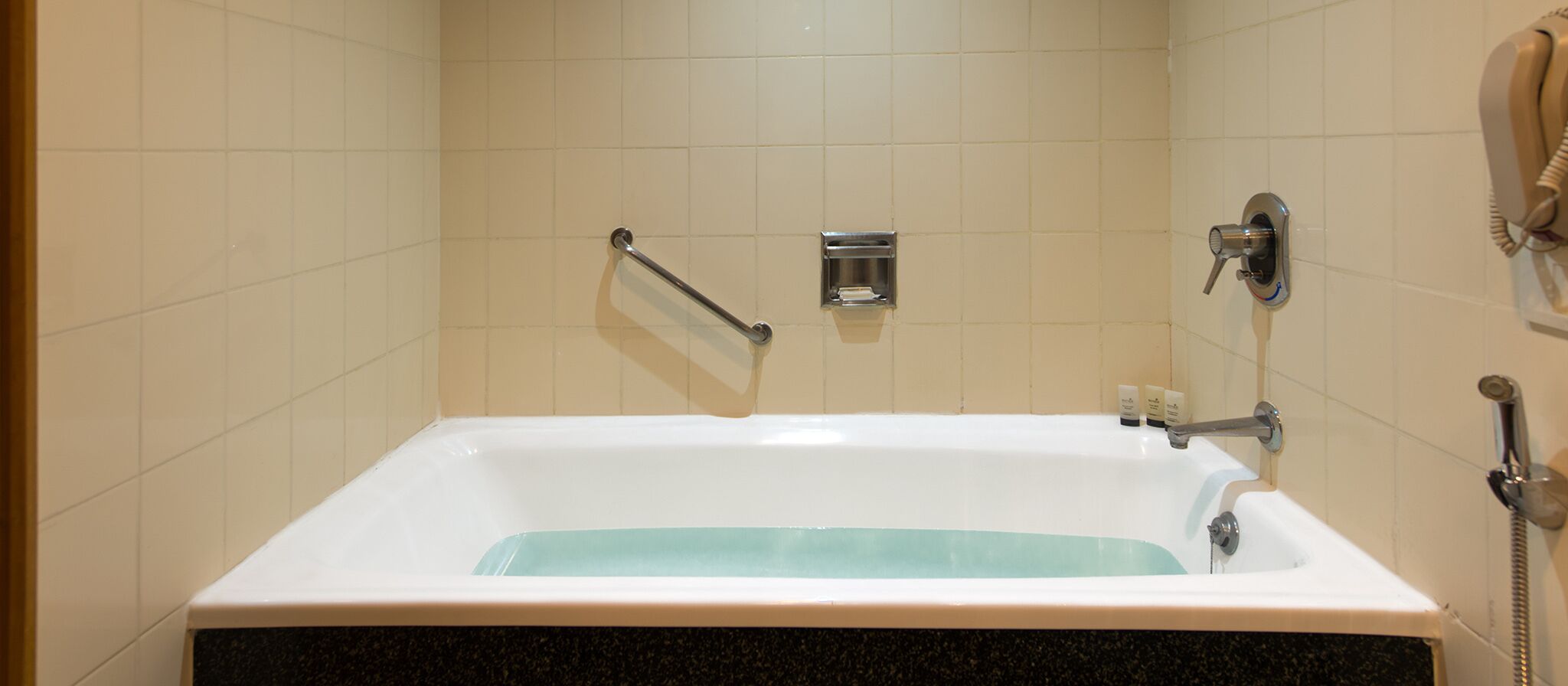 Una bañera con un cabezal de ducha manual, un perchero y un dispensador de jabón en la pared.