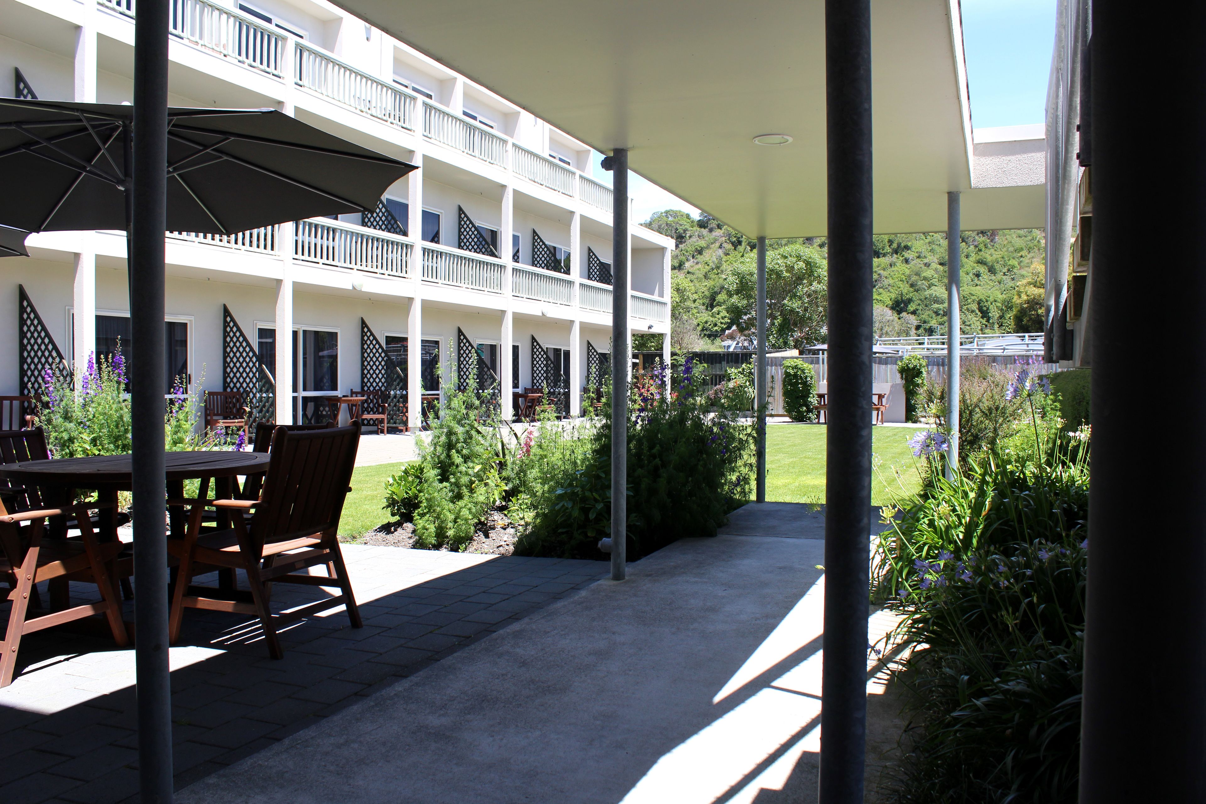 A covered outdoor seating area with a table and chairs, surrounded by greenery and a white building with balconies.