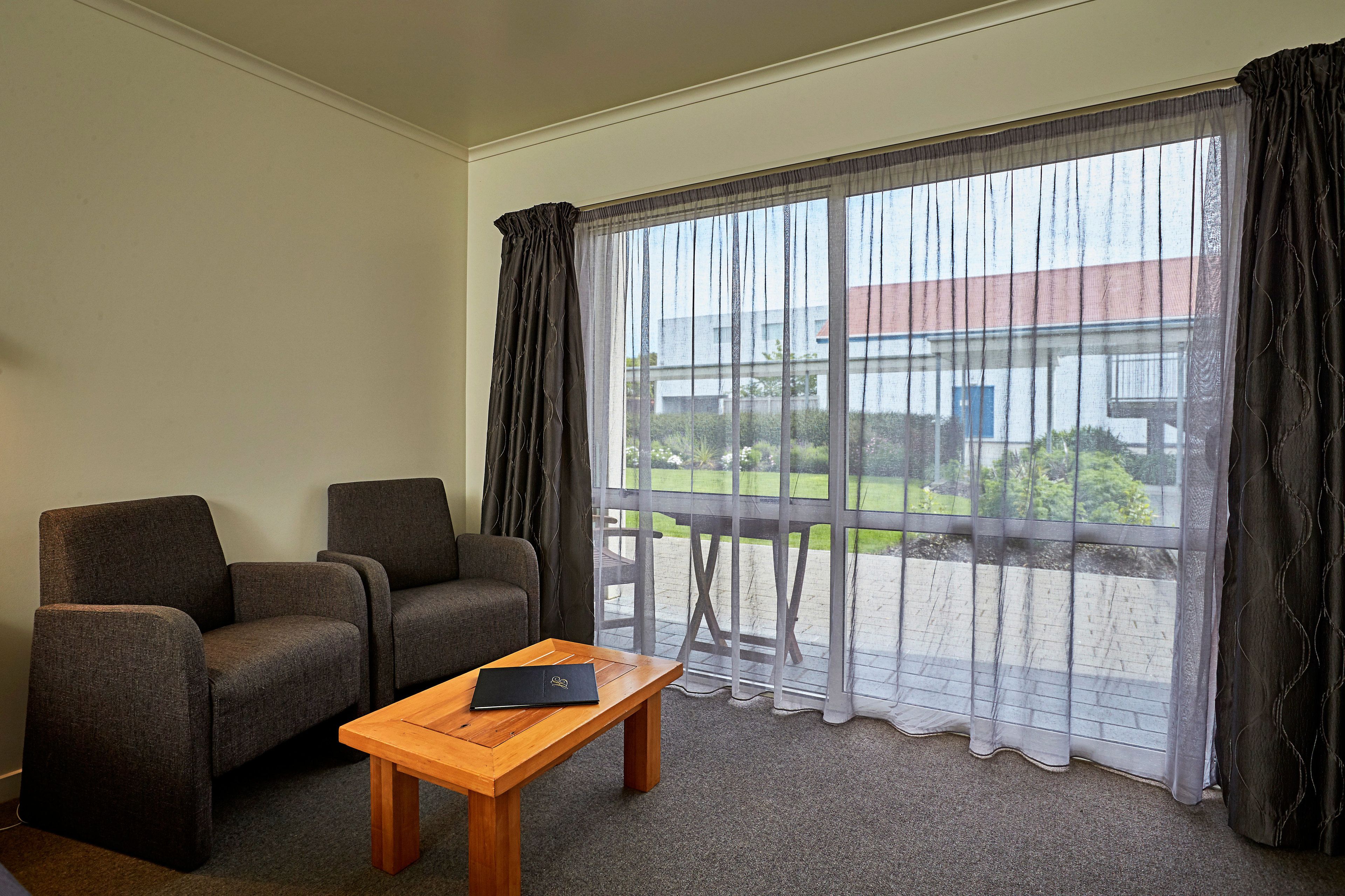 A room with two grey armchairs, a wooden coffee table with a laptop, and a large window with sheer curtains.