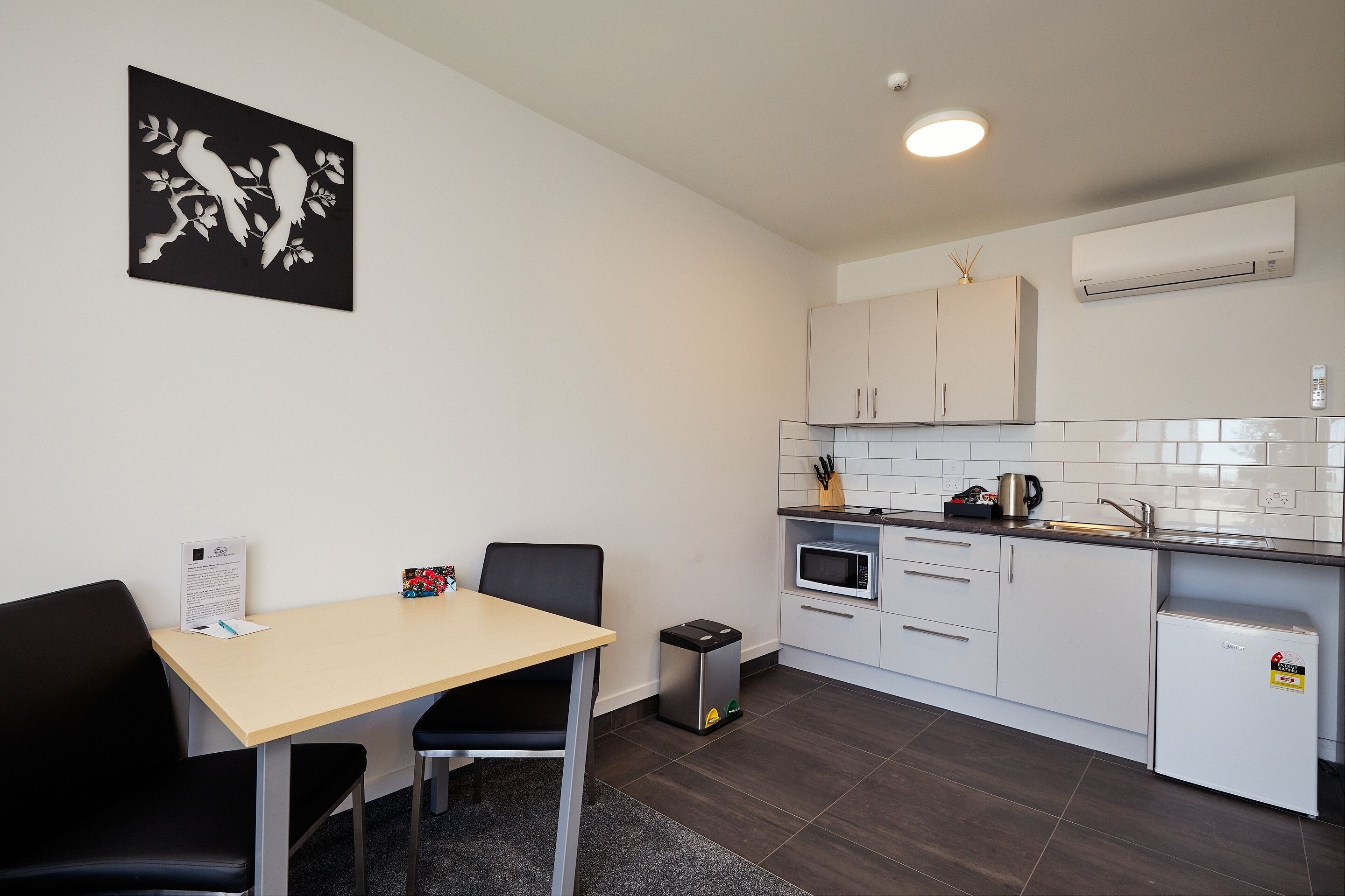 A compact kitchen with white cabinets, a microwave, and a small refrigerator. There is a black chair and a table with a menu on it.