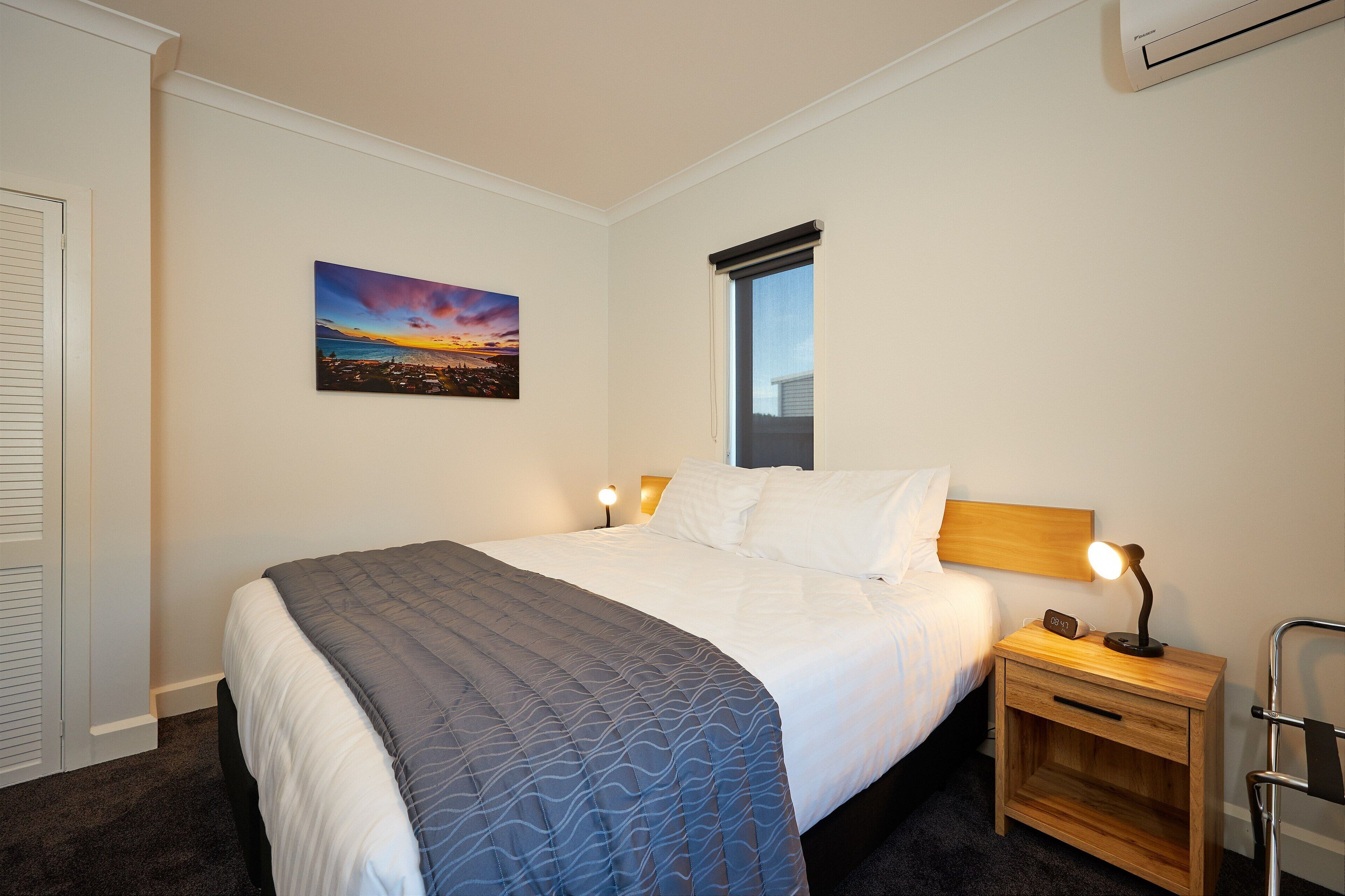 A neatly made bed with a blue and white bedspread, a nightstand with a lamp, and a painting of a sunset on the wall.