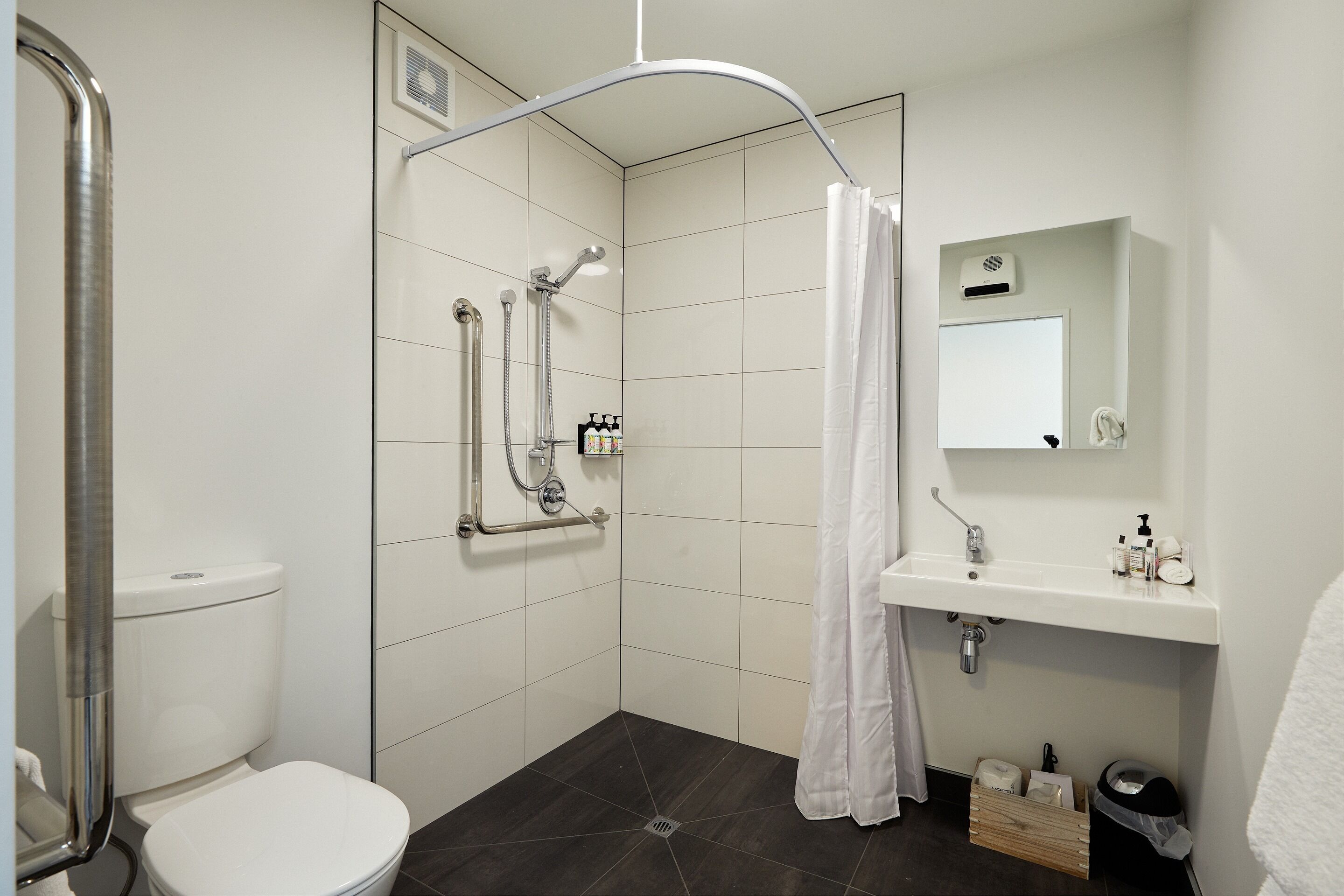Ein modernes Badezimmer mit Toilette, einer Dusche mit gebogener Duschbrause, einem weißen Waschbecken mit Spiegel darüber und einer kleinen Mülleimer.