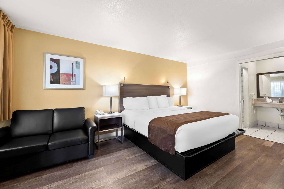 A hotel room with a bed, a nightstand, a black leather sofa, a painting on the wall, and a bathroom visible through an open door.