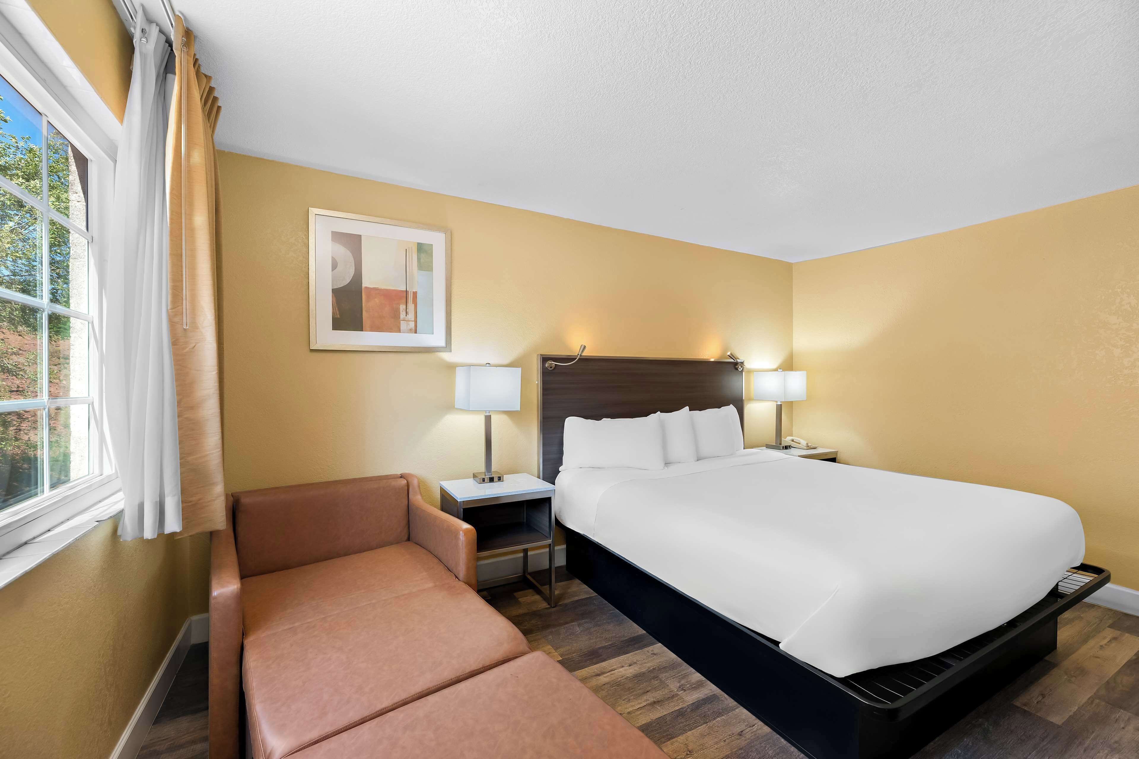 A hotel room with a large bed, a sofa, a nightstand with a lamp, and a painting on the wall.