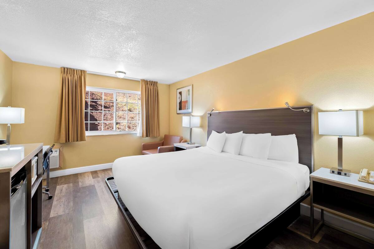 A hotel room with a large bed, a desk with a lamp, a chair, and a window with curtains.
