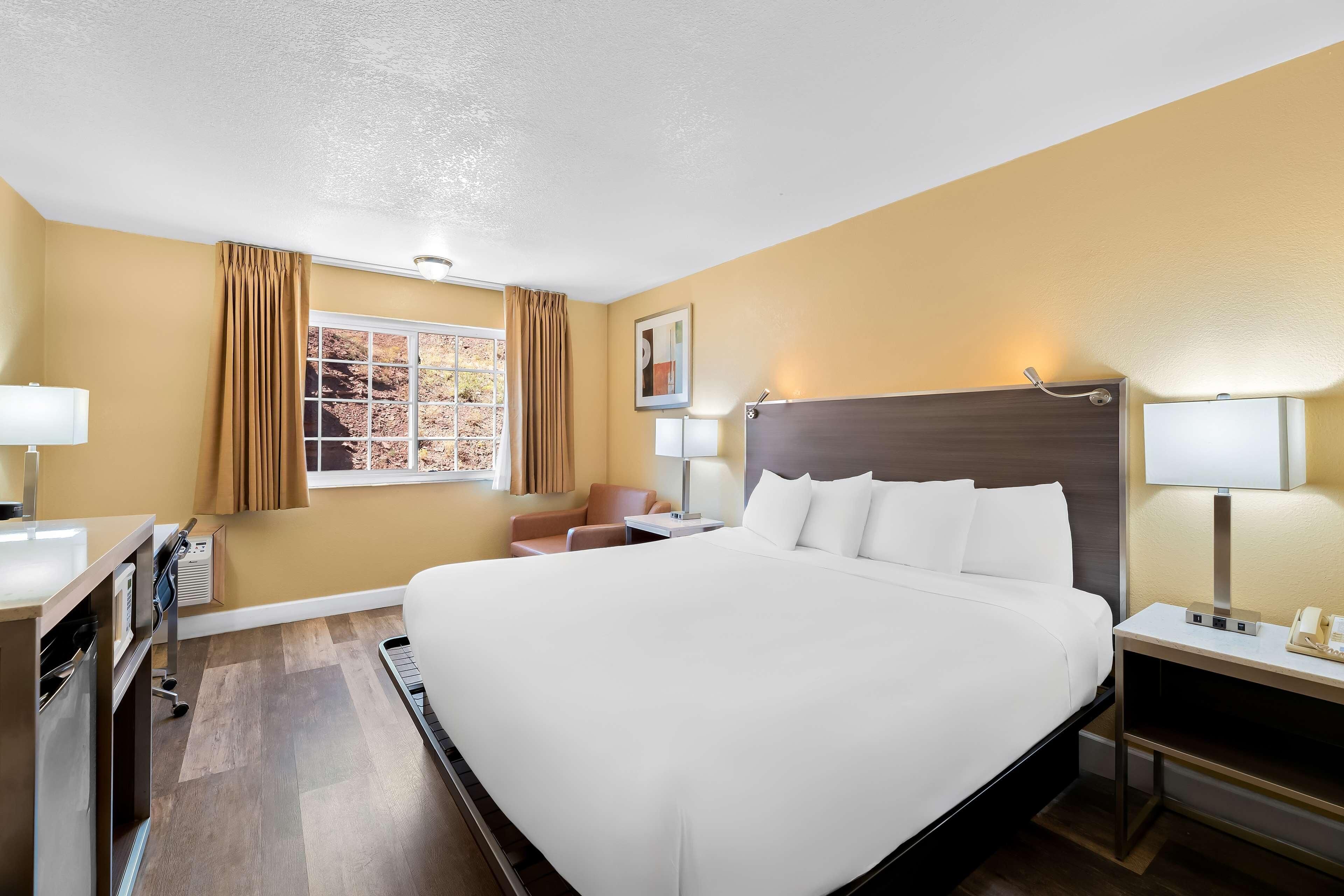 A hotel room with a large bed, a desk with a lamp, a chair, a window with curtains, and a painting on the wall.