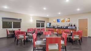 A restaurant with multiple tables and chairs, a counter with food and drink options, and a door on the right.
