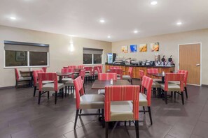 A restaurant with multiple tables and chairs, a counter with food and drink options, and a door on the right.
