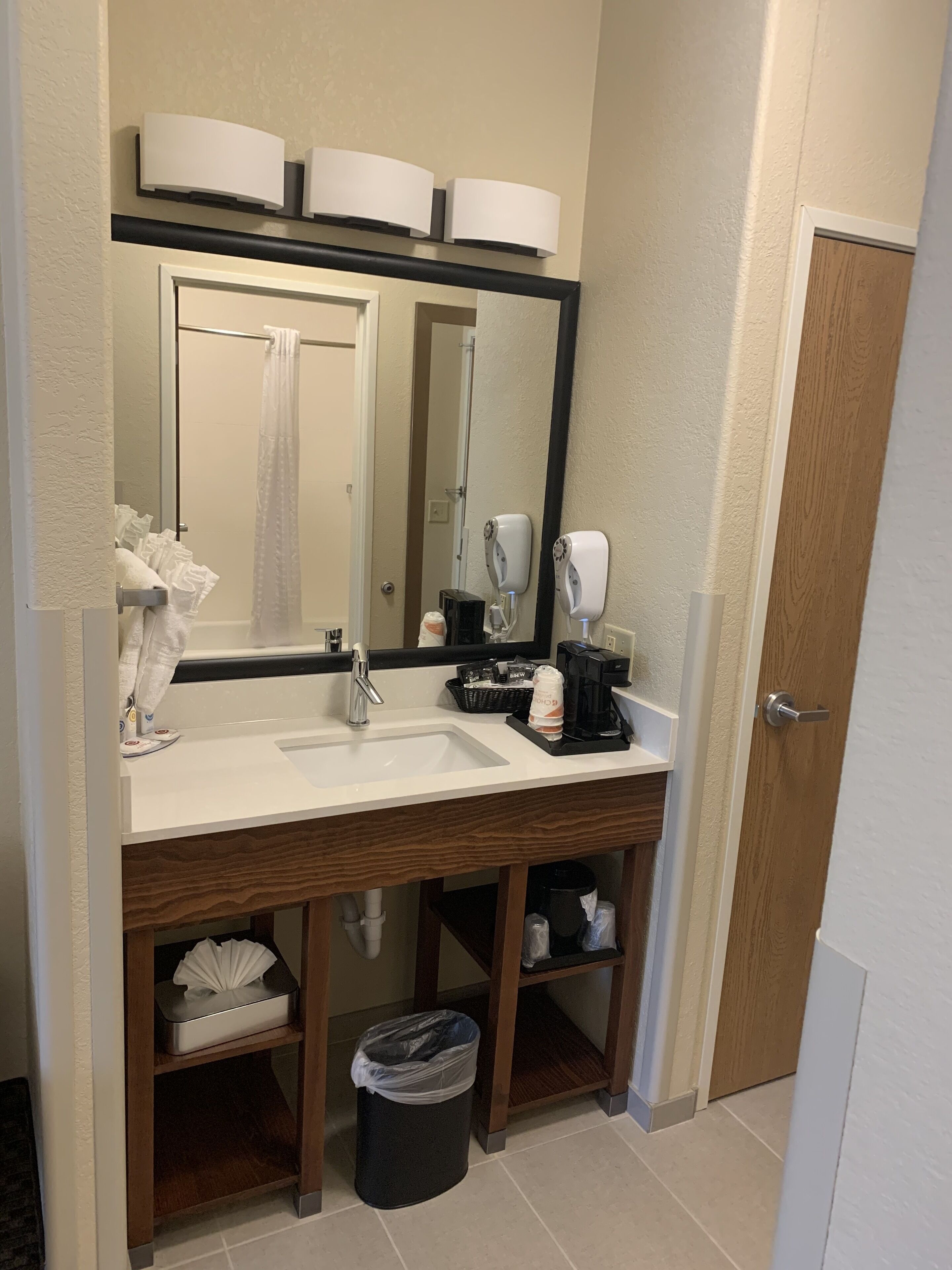 A bathroom with a white sink, a mirror, a towel rack, a trash can, and a coffee maker.