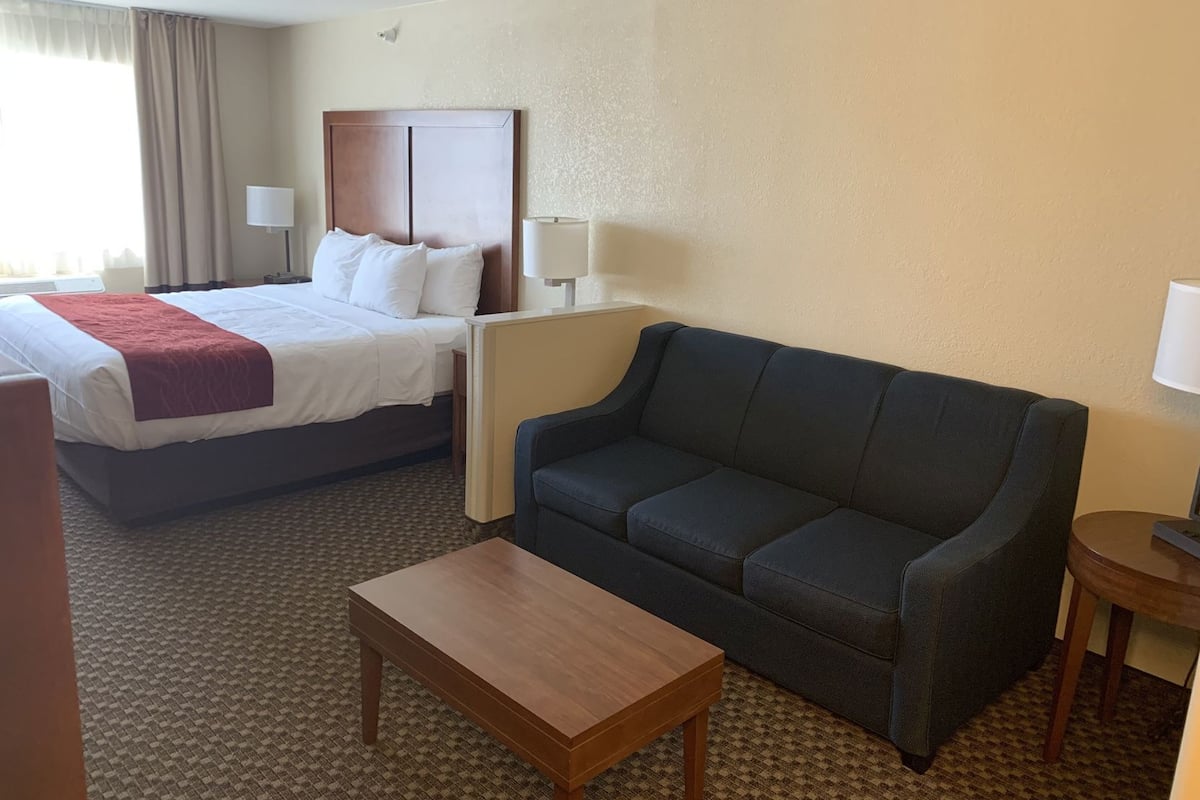 A hotel room with a bed, a sofa, a small table, and a nightstand with a lamp.