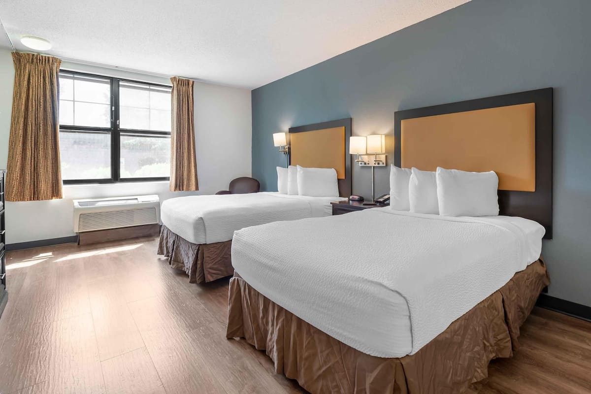 A hotel room with two beds, a large window with curtains, a radiator, and a wall-mounted lamp.