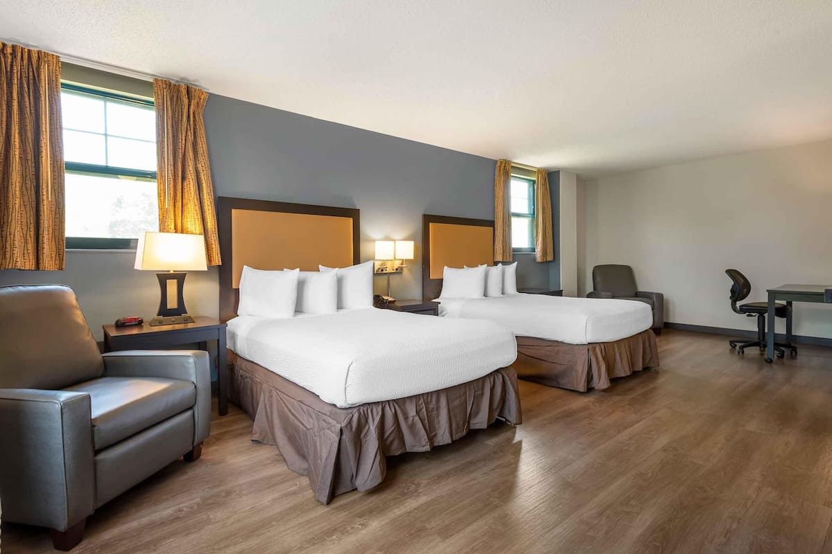 A hotel room with two beds, a desk, a chair, and a lamp.