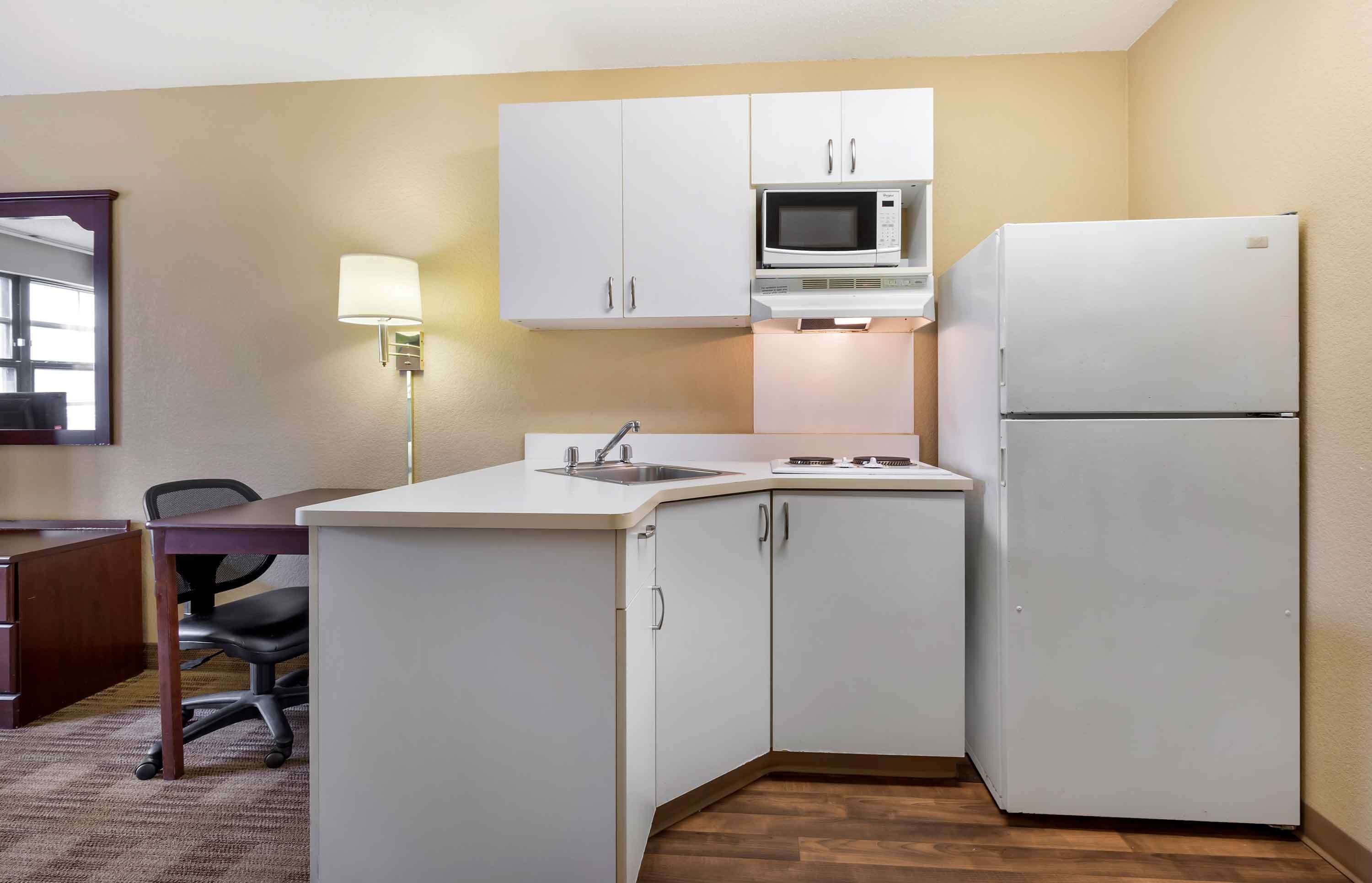 A compact kitchen with white cabinets, a microwave, a refrigerator, a sink, and a desk with a chair.