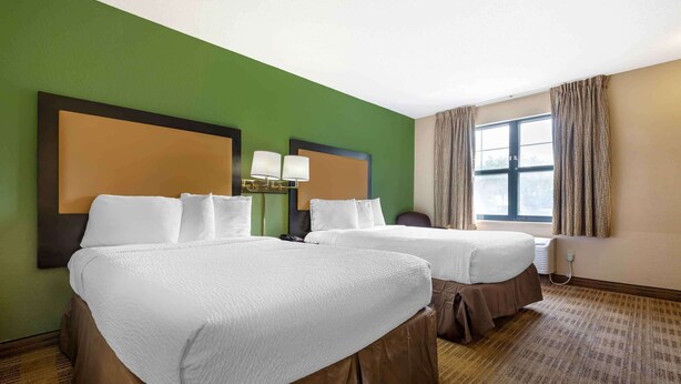 A hotel room with two beds, a green wall, a large window with curtains, and a headboard with lamps.