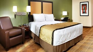Ein Hotelzimmer mit Bett, Sessel, Nachttisch, Lampe und einem gerahmten Bild an der Wand.