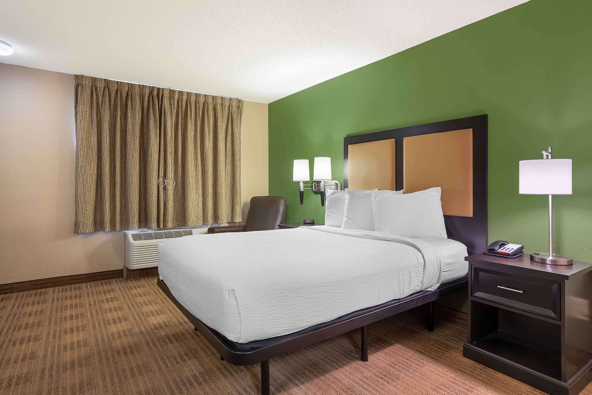 Habitación de hotel con cama, mesita de noche, lámpara, silla y un ventanal con cortinas.