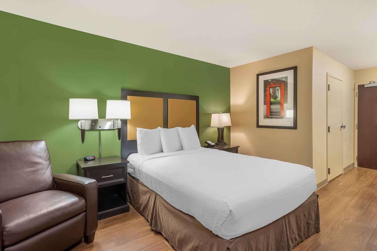 Una habitación de hotel con cama, mesita de noche, lámpara, silla y puerta.