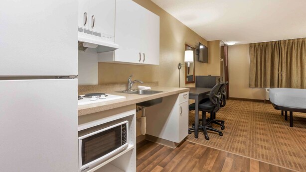A compact kitchen with a microwave, oven, sink, and cabinets. There is a dining area with a table and chairs, and a large window with curtains. - Extended Stay America Select Suites - Columbia - Stadium Blvd. (Columbia)